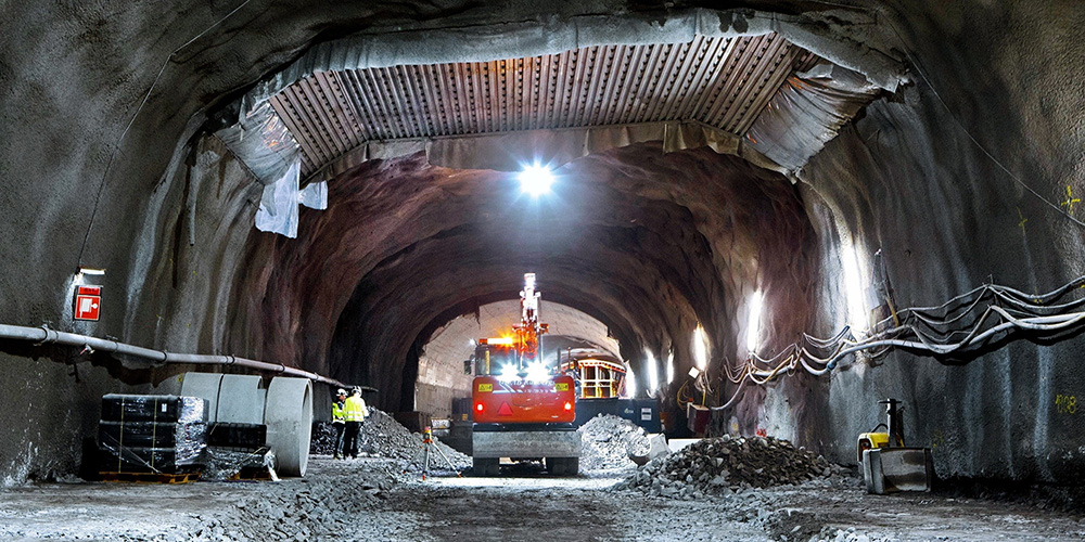Arbetsmaskin och två varselklädda personer i tunnel under uppbyggnad.