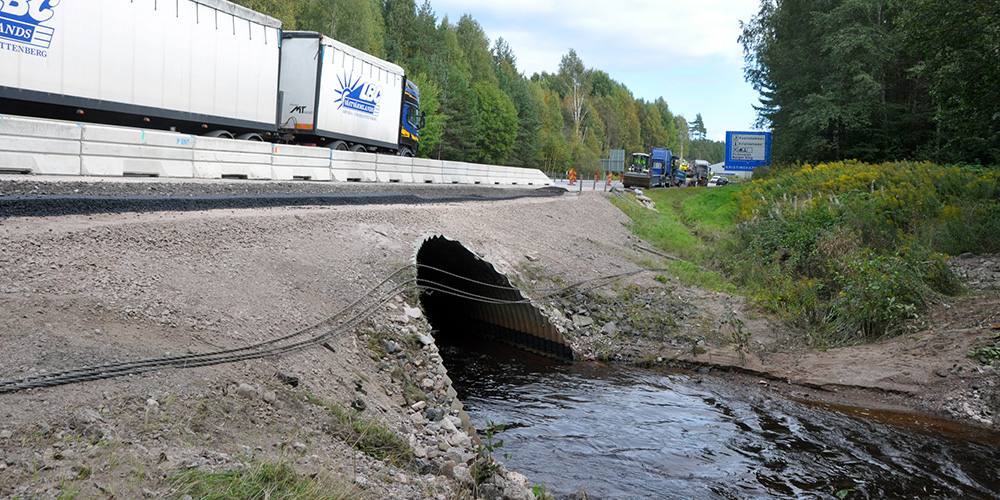 Vattendrag rinner genom vägtrumma under trafikerad väg.