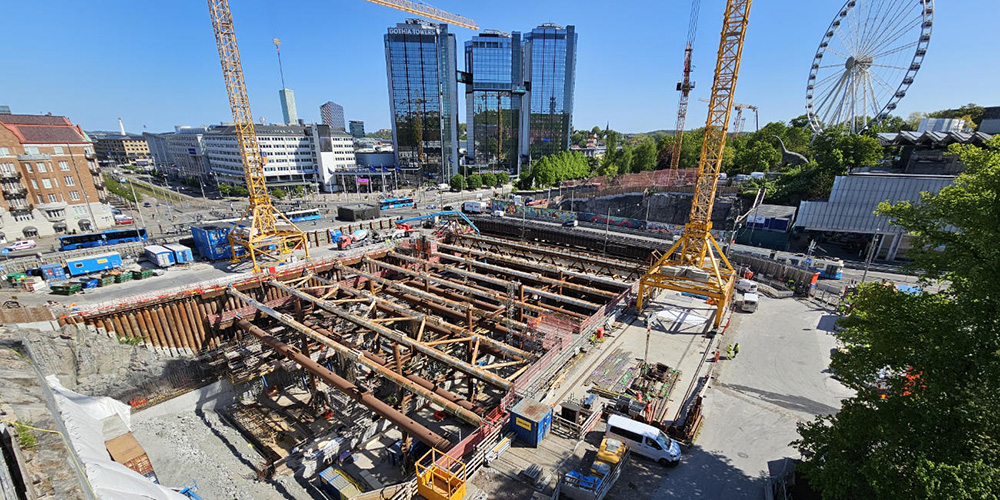 Arbetsområde med stålbalkar och lyftkranar. I bakgrunden syns stad och nöjesfält.