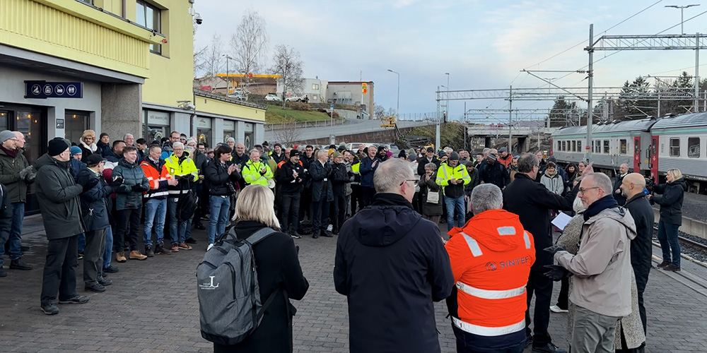 En folksamling mellan en gul byggnad och ett spårområde.