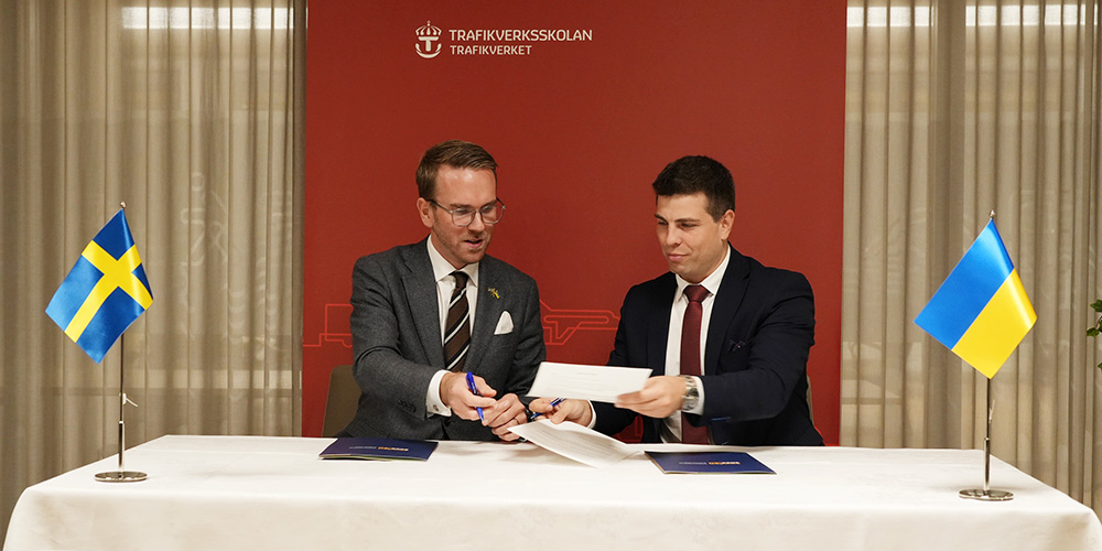 Två män delar papper vid ett bord med Sveriges och Ukrainas flaggor.