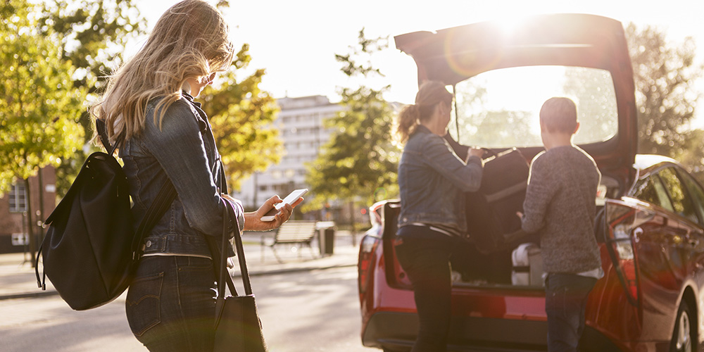 En kvinna och en pojke packar en bil medan en ung kvinna tittar i en mobil.