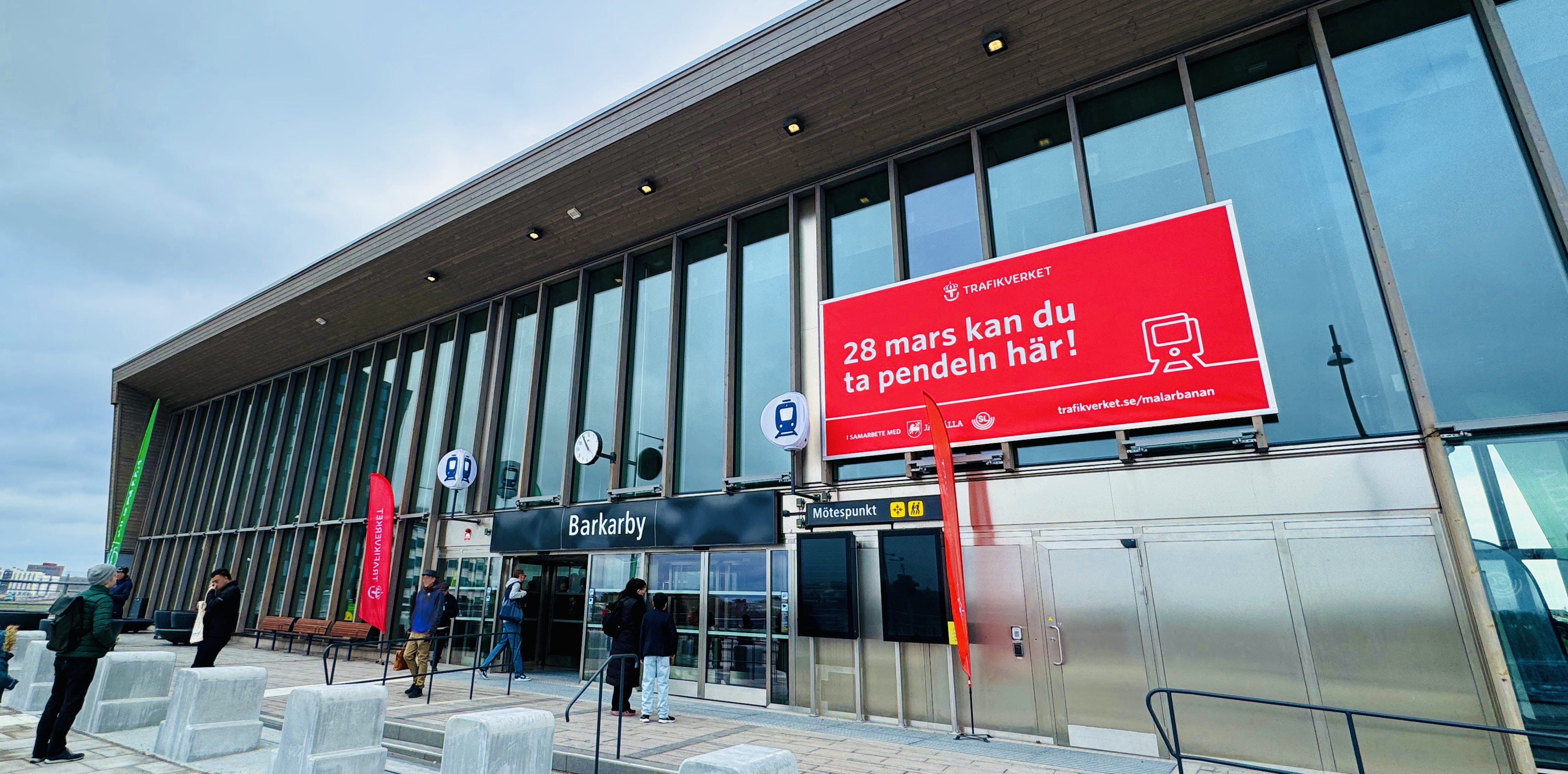 husfasad med stora glasfönster med röd vepa med vit text Barkarby öppnar 28 mars